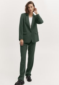 Zaļš pielāgots uzvalks ar vienu pogu un taisnu galu biksēm. Audums ir ar gludu tekstūru, kas papildināts ar baltu topu zem tā.