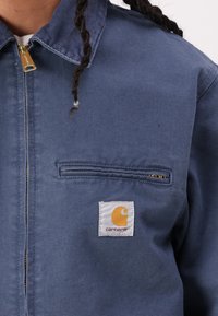 Jachetă din denim albastru cu fermoar frontal, un buzunar pe piept și un eticheta pătrată albă Carhartt cu un emblema galbenă.