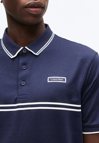 Άνδρας που φοράει μπλε σκούρο πόλο με λευκές διπλές ρίγες και το λογότυπο Calvin Klein στο στήθος, κοντινή λήψη από τους ώμους μέχρι τα χείλη.