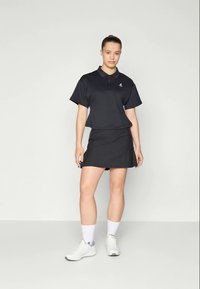 Juodas polo marškinėliai su apykakle ir trumpomis rankovėmis, derinami su juoda sijonu. Lygi tekstūra, minimalistinis dizainas ir baltos sportinės bateliai.