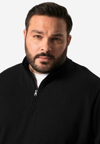 Sudadera negra con cremallera hecha de una tela suave, con un cuello alto y una cremallera de un cuarto; presenta una camisa blanca debajo.