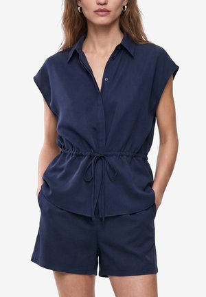 Vrouw draagt marineblauwe mouwloze top met kraag en trekkoord in de taille en bijpassende short, handen in de zakken, tegen een effen achtergrond.