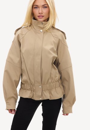 Femme aux cheveux blonds portant une veste beige à col montant avec taille élastique et un pantalon noir sur un fond uni.