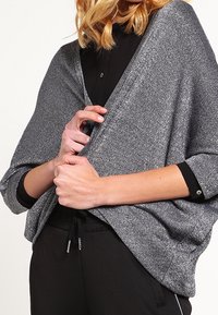 Metalický šedý šál s volným střihem, měkkou texturou a širokými rukávy, nošený přes černou košili a doplněný černými tepláky.