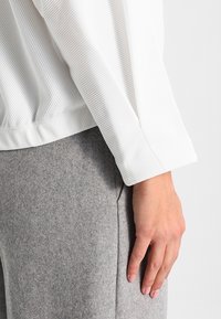 Hand resting by side wearing a long-sleeve white ribbed top and gray woolen pants, shown from waist to mid-thigh.