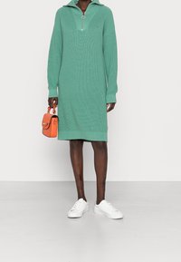 Robe pull en maille verte avec un col zippé, des manches longues et une coupe droite. Associée à un sac à main orange et des baskets blanches.