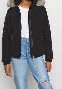 Veste matelassée noire avec une capuche en fourrure amovible, fermeture éclair à l'avant et logo ton sur ton. Associée à un jean déchiré bleu clair.