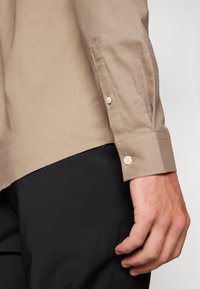 Camicia beige a maniche lunghe con una texture liscia, polsini con bottoni e vestibilità strutturata, abbinata a pantaloni neri aderenti.