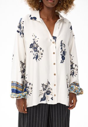 Kobieta w oversize białej koszuli zapinanej na guziki z niebieskimi kwiatowymi wzorami i szerokimi mankietami w niebiesko-żółte wzory, zestawiona z ciemnymi spodniami w cienkie prążki.
