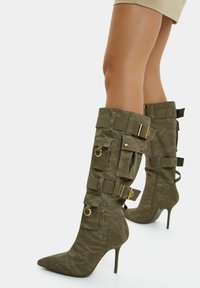 Olivgröna knähöga stövlar med texturerad finish, spetsig tå, hög stilettklack och dekorativa detaljer inklusive spännen och fickor.