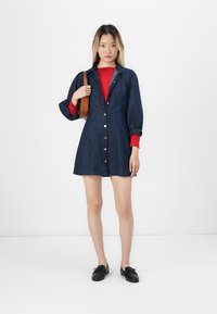 Jeansklänning med knäppning framtill, figurnära liv, puffärmar och A-linjeformad kjol. Bärs över en röd topp, tillsammans med svarta skor och en brun handväska.