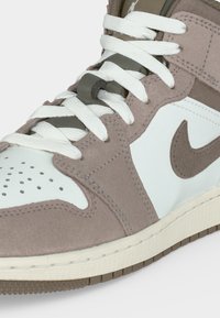 Κοντινό πλάνο σε sneaker Nike σε απόχρωση ταούπ και λευκό, εστιάζοντας στα κορδόνια, τα δερμάτινα και καστόρινα πάνελ, το διάτρητο κουτί στα δάχτυλα και το ορατό λογότυπο Nike swoosh.