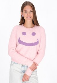 Rosa Pullover mit einer flauschigen Textur, versehen mit einem großen lila Smiley-Gesichtdesign. Gerippte Bündchen und Saum, kombiniert mit hellen Jeans.