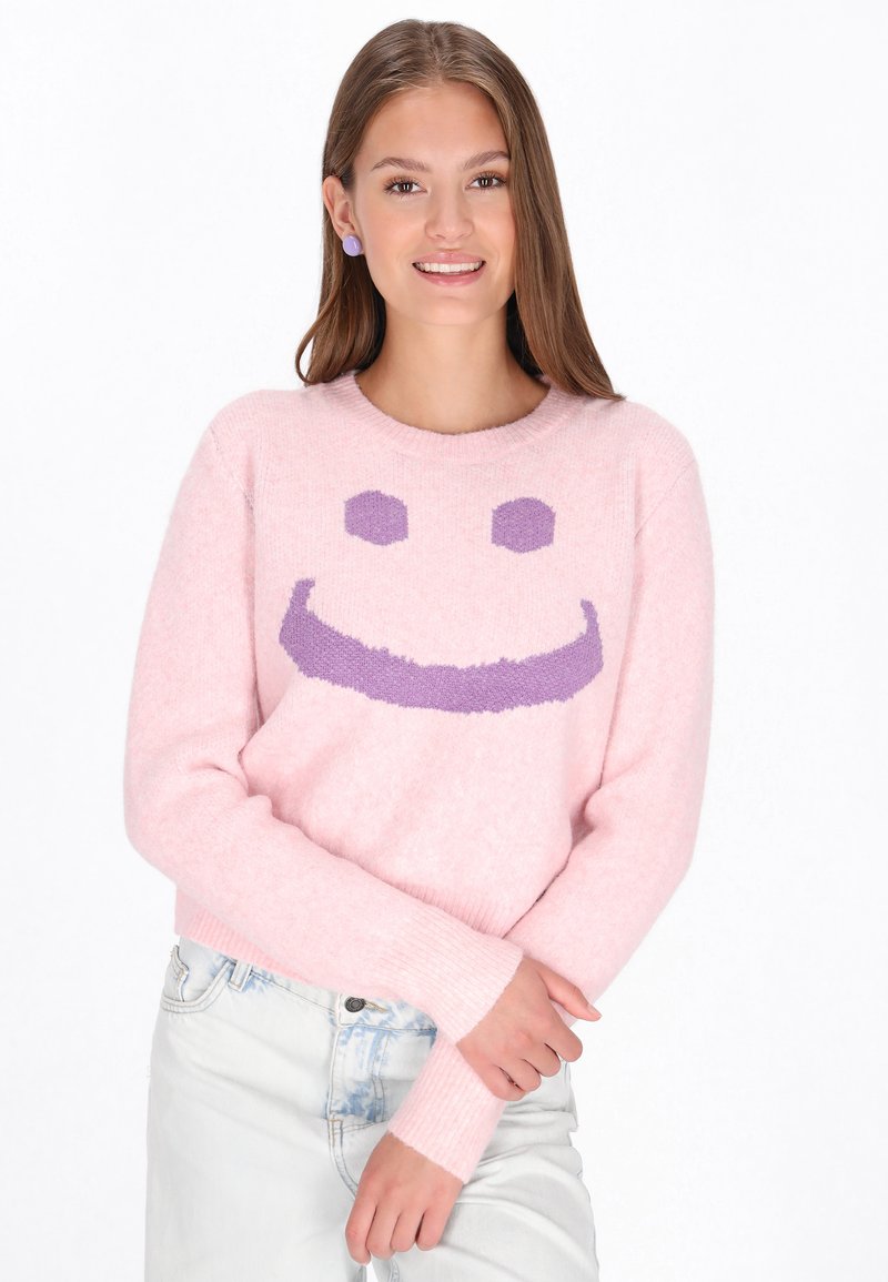 Rosa Pullover mit einer flauschigen Textur, versehen mit einem großen lila Smiley-Gesichtdesign. Gerippte Bündchen und Saum, kombiniert mit hellen Jeans.