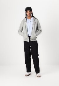 Hanorac gri cu fermoar peste un tricou bleu deschis, pantaloni cargo negri, șapcă neagră și adidași albi cu accente maronii; caracteristici includ buzunare și o croială confortabilă.