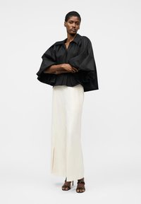 Femme debout, les bras croisés, portant un haut noir à manches volumineuses, un pantalon large crème avec des fentes à l'avant, et des escarpins noirs à bouts ouverts.