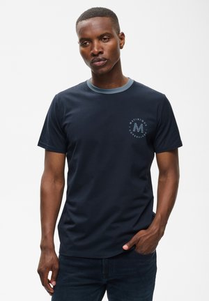 Mand iført en mørkeblå kortærmet t-shirt med en lyseblå krave og et lille rundt logo på venstre bryst, står med den ene hånd i lommen.