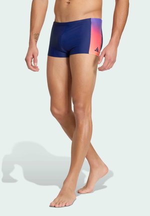 Mann trägt marineblaue Badehose mit pinkem Farbverlauf an der Seitenpartie, steht barfuß vor einem blassen Hintergrund.