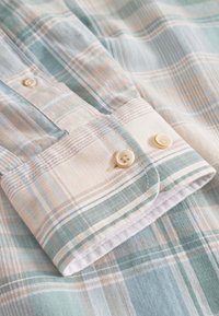 Camicia a quadri leggeri realizzata in morbido cotone, con colori blu, verde e beige. Presenta un polsino con bottoni e cuciture dettagliate.