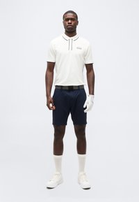 Άνδρας που στέκεται, φορώντας λευκό πόλο, μπλε σκούρες σορτς, λευκό γάντι του γκολφ στο αριστερό χέρι, λευκές κάλτσες και λευκά αθλητικά παπούτσια, μπροστά σε απλό φόντο.