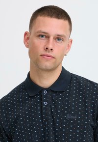 Polo noir avec un motif de carrés bleu clair et blancs, deux boutons, et un logo brodé "Blond" sur la poitrine.