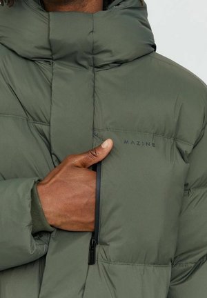 Main fermant la poche poitrine zippée sur une doudoune verte de la marque Mazine avec un col montant.