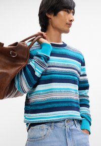 Un pull rayé avec des nuances de bleu variées, présentant des motifs texturés et des poignets côtelés. Associé à un jean bleu clair et un sac en cuir marron.
