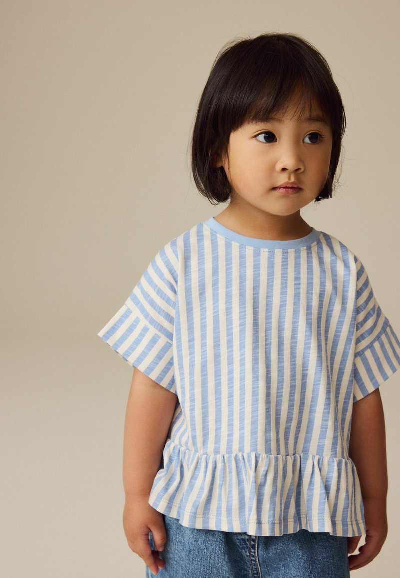 Bambino piccolo con capelli neri corti che indossa una camicia a righe verticali blu e bianca con orlo a volant e pantaloni di jeans, che guarda di lato.