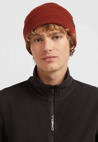 Gorro de punto rojo con textura acanalada, llevado por una persona con una chaqueta de forro polar negra con cremallera y letras de marca blancas. Vista en primer plano.