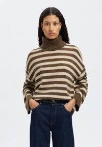 Ränderad tröja i ljusbeige och brun, tillverkad av ett mjukt material. Har en hög polokrage och vida ärmar. Bärs med mörkblå jeans.