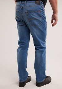 Mees, kes kannab siniseid teksapükse ja musti kingi, seisab käsi vabal küljel ühetaolise tausta ees.