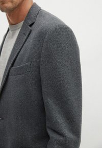 Uomo che indossa un blazer grigio a spina di pesce sopra una shirt a girocollo grigia chiara, accorciata al collo e alle maniche.