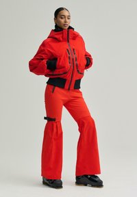 Femeie în picioare cu mâinile în buzunare, purtând o jachetă roșie cu fermoar, pantaloni evazați asortați cu cataramă neagră și bocanci de schi negri.