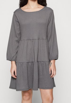 Kvinde iført en grå, langærmet, lagdelt kjole med elastiske manchetter, stående mod en enkel lys baggrund.