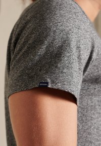 Close-up van de bovenarm van een persoon die een kortdurige, grijze Superdry t-shirt draagt, waarbij de textuur van de stof en het merklabel zichtbaar zijn.