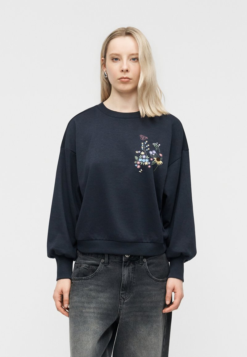 Ξανθιά γυναίκα που φορά υπερμεγέθη μαύρο φούτερ με πολύχρωμη κεντητή λουλουδάτη διακόσμηση στο στήθος και ξεβαμμένο μαύρο τζιν, μπροστά σε απλό φόντο.