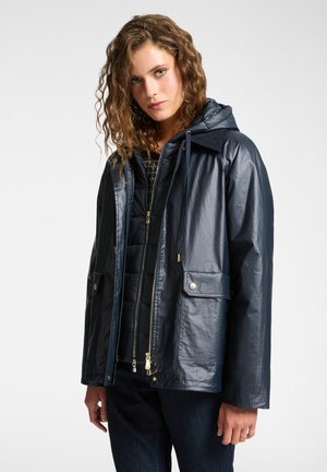 Chaqueta impermeable de color navy con capucha, cierre delantero con cremallera, dos bolsillos frontales y detalles de hardware dorado. Acabado texturizado y corte relajado.