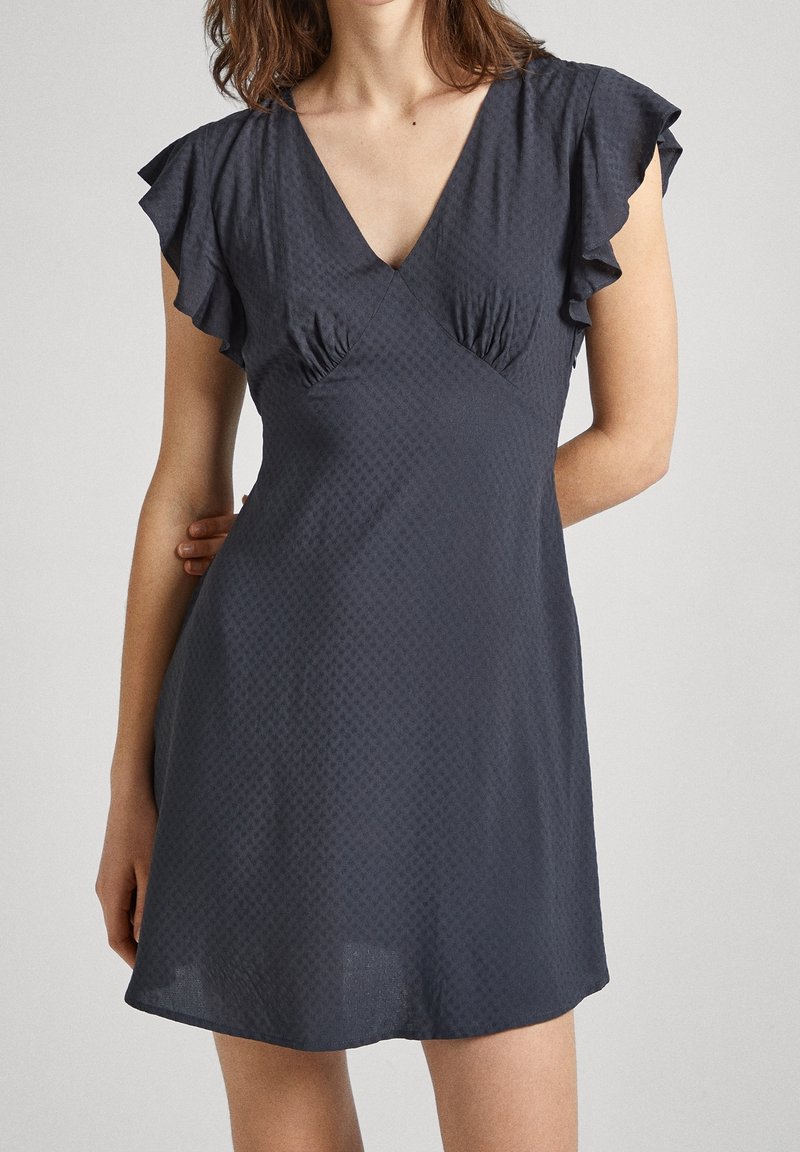 Femme portant une robe courte gris foncé avec des manches volantées et un col en V, debout devant un fond clair uni.