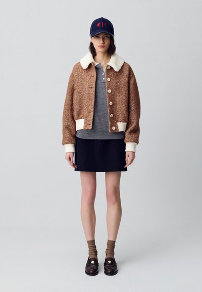 Chaqueta marrón de espiga con cuello de piel blanca, camisa gris debajo, falda negra, calcetines marrones y mocasines oscuros con detalles en dorado.