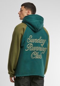 Kapucnis pulóver zöld és olíva panelekből. A hátán sárga hímzésű "Sunday Running Club" felirattal. Puha anyag, laza szabású.