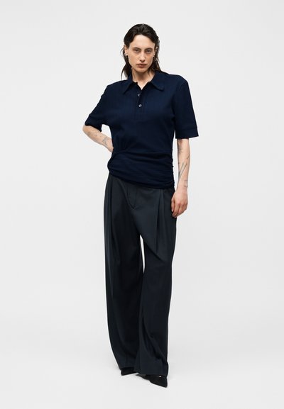 Άτομο με τατουάζ που φοράει μπλε ναυτικό κοντομάνικο πουκάμισο με γιακά και φαρδιά μαύρη παντελόνια, στέκεται μπροστά σε απλό φωτεινό φόντο.