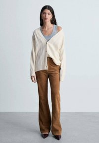 Vaaleanbeige neulecardigan, jossa on V-pääntie ja napit, päällä harmaa toppi. Muotoiltu ruskea mokkapantalon, jossa on leveät lahkeet, ja burgundinpunaiset korot.