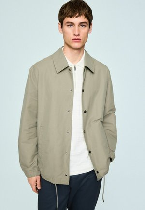 Young man wearing a beige button-up jacket over a white zip-neck shirt and dark pants, standing against a plain light background.