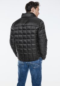 Černá péřová bunda s prošívaným vzorem, vysokým límcem a lesklou texturou, nošena s modrými džíny. Zadní pohled ukazuje obepínající tvar.