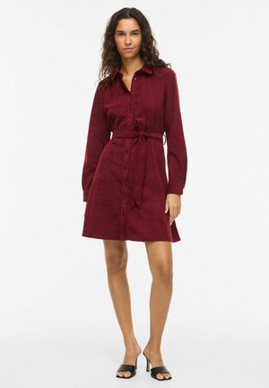 Robe-chemise en velours côtelé bordeaux avec manches longues, boutonnée sur le devant et taille ceinturée ; texture lisse et forme trapèze. Des sandales complètent le look.