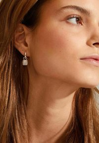 Boucles d'oreilles pendantes en argent avec un charme en forme de cadenas texturé, doté d'un attache en anneau lisse, portées sur l'oreille d'un modèle.