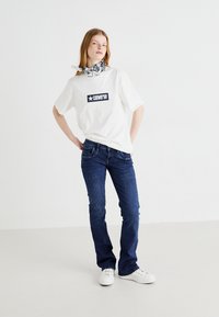 Biela tričko s námorníckym logom "Converse", spárované s tmavomodrými džínami a bielymi teniskami. Model má na sebe šatku s čierno-bielym vzorom.