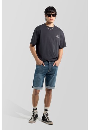 Giovane uomo con occhiali da sole neri, t-shirt nera oversize, pantaloncini di jeans rimboccati, calzini a righe e sneakers nere alte.