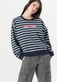 Marineblå og hvite stripete genser med rød brodert "NEW YORK" tekst, rund hals og ribbet mansjetter og hem. Bært over grå bukser.