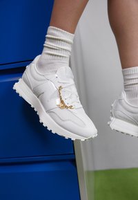 Weiße Sportsneaker aus Mesh und synthetischen Materialien, akzentuiert mit einem goldenen Kettendetail, auf einem Bein mit gerippten cremefarbenen Socken präsentiert.